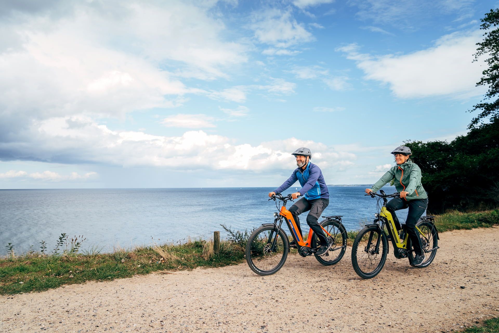 Zwei Personen fahren mit E-Bikes auf einem Schotterweg entlang eines Gewässers unter bewölktem Himmel
